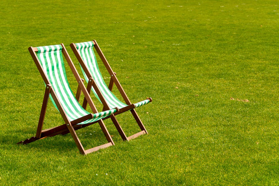 Two wooden deck chairs with green and white striped fabric seats and backs are positioned on a lush, well-maintained lawn. The grass is vibrant, evenly cut, and slightly textured, with a few small patches of dry soil or scattered leaves visible in the background. The garden features a border of neatly trimmed hedges and a pathway that runs along the edge of the lawn. The scene is evenly lit, suggesting daylight, and conveys a calm outdoor environment suitable for relaxation or gardening activities. The image is associated with Gardeners Crystal Palace, highlighting garden maintenance and landscaping work relevant to lawn care and outdoor leisure spaces.