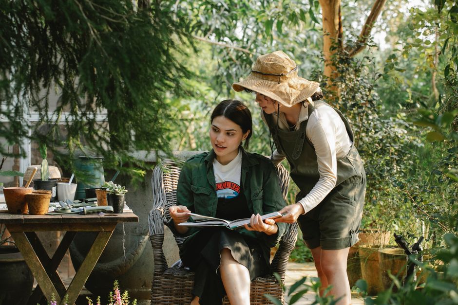 A woman and a young girl are situated in a lush garden setting, surrounded by green foliage, trees, and potted plants on a wooden outdoor table. The woman, wearing a wide-brimmed straw hat, sunglasses, and a long-sleeve shirt under a green apron, leans over to show the girl a book, engaging in garden maintenance or landscaping advice. The girl, seated on a wicker chair, has dark hair tied back, and is dressed casually in a jacket and skirt, attentively looking at the book. The garden's natural light filters through the leaves, illuminating the scene with a warm, inviting atmosphere. The background includes various gardening tools, containers, and ongoing plant care activities, characteristic of a well-tended outdoor space maintained by local gardeners such as Gardeners Crystal Palace, as highlighted in the Westow Hill Crystal Palace garden care guide.